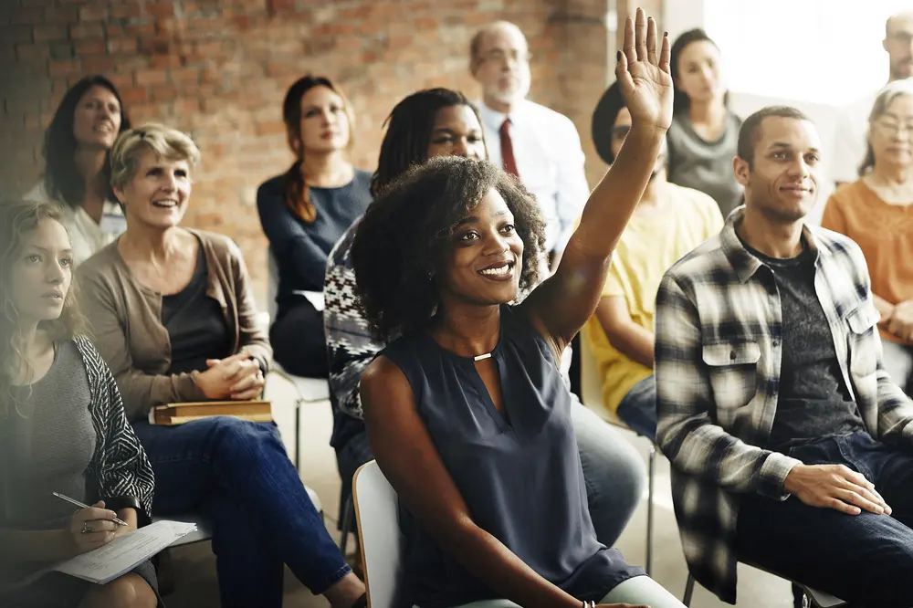 Black woman smiling while raising her hand to ask a question at a conference presentation